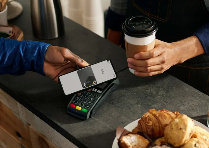 Eine Person nutzt ein Smartphone für eine kontaktlose Zahlung an der Café-Theke. Zu sehen sind ein Bezahlterminal und verschiedene Backwaren in der Auslage. Eine weitere Person hält eine Kaffeetasse in entspannter Atmosphäre. Auf dem Smartphone ist eine Bezahloberfläche mit sichtbaren Symbolen zu sehen.