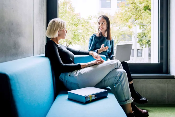 Zwei Frauen sitzen auf einem blauen Sofa vor einem großen Fenster und führen ein lebhaftes Gespräch. Eine der Frauen hält einen Laptop, während in der Nähe ein Aktenordner liegt – ein Hinweis auf eine produktive Zusammenarbeit oder eine gemeinsame Lerneinheit in einer hellen, modernen Umgebung.