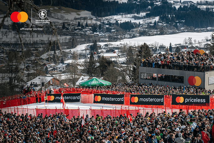 Eine große Menschenmenge versammelt sich bei einer Outdoor-Wintersportveranstaltung, im Hintergrund schneebedeckte Hügel und Bäume. Die Szene zeigt eine Zuschauerplattform mit Mastercard-Branding und dem gut sichtbaren Schriftzug „priceless“. Logos von Mastercard und „Official Partner“ sind oben links zu sehen.