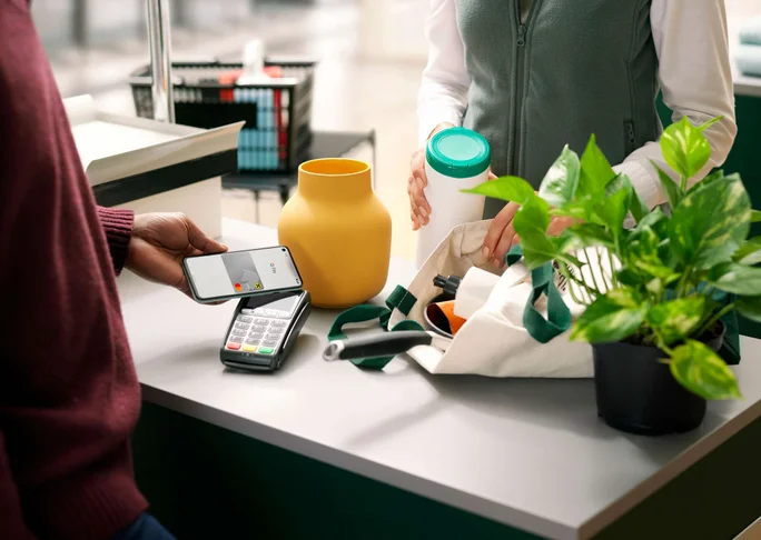 Eine Person nutzt ein Smartphone für eine kontaktlose Zahlung an der Kasse. Die Umgebung wirkt wie ein nachhaltiges Geschäft mit wiederverwendbaren Artikeln wie einer gelben Vase und einer Stofftasche. Das Bezahlterminal zeigt einen Transaktionsbildschirm, und eine grüne Topfpflanze unterstreicht das nachhaltige Thema. Die Szene hebt moderne Technologie und bewusstes Einkaufsverhalten hervor.