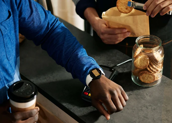 Eine Person mit Smartwatch tätigt eine kontaktlose Zahlung an der Theke einer Bäckerei. Im Setting sind ein Glas mit Keksen und eine Papiertüte zu sehen, die von einer weiteren Person befüllt wird. Die Szene betont moderne Technologie in einem alltäglichen, entspannten Umfeld mit warmen Farbtönen und natürlichem Licht.