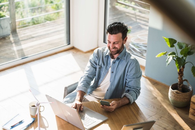 Ein Mann sitzt an einem Holztisch in einem sonnendurchfluteten Homeoffice und nutzt ein Smartphone, während vor ihm ein Laptop geöffnet ist. Die Umgebung ist geprägt von großen Fenstern, viel Tageslicht und Zimmerpflanzen, was eine entspannte und produktive Atmosphäre schafft. Auf dem Schreibtisch stehen außerdem eine Kaffeetasse und ein Notizbuch, was das Arbeitsumfeld abrundet.